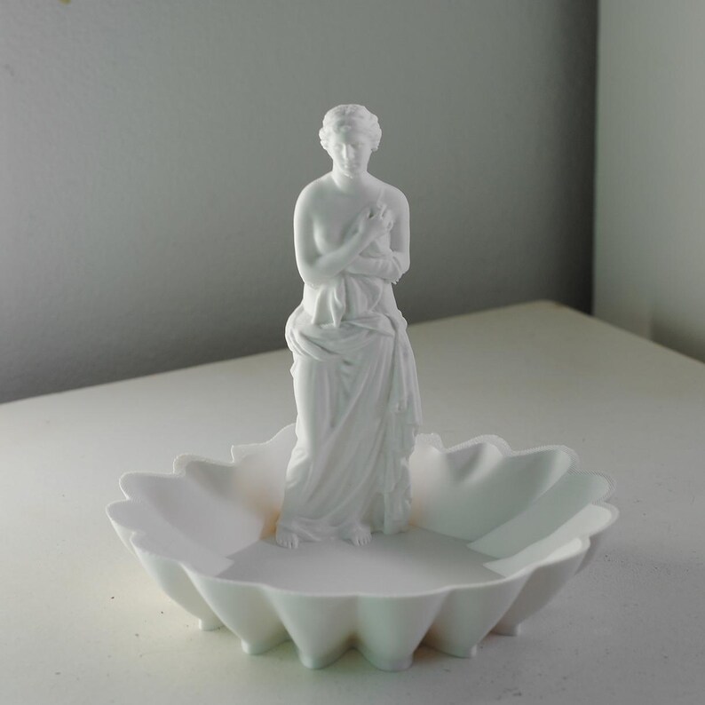 Puede incluir: Una bandeja decorativa de color blanco mate que presenta una figura central de una mujer de pie envuelta en tela. La base de la bandeja tiene forma de concha festoneada con crestas profundas y redondeadas que irradian desde el centro.