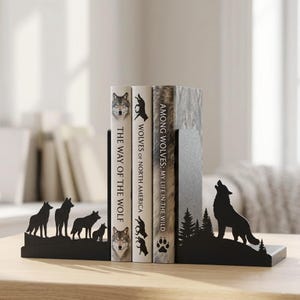 Puede incluir: Un par de sujetalibros de metal negro con siluetas de lobos. La pieza izquierda muestra una manada de lobos, mientras que la derecha representa un lobo aullando frente a pinos. Tres libros est&aacute;n colocados entre los dos soportes met&aacute;licos.