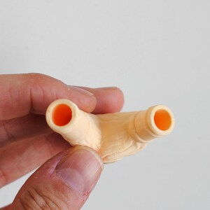 Puede incluir: Un componente de pl&aacute;stico de color melocot&oacute;n claro con una estructura en forma de Y y dos aberturas cil&iacute;ndricas huecas en la parte superior. La superficie muestra un acabado fino y texturizado, caracter&iacute;stico de la fabricaci&oacute;n 3D.