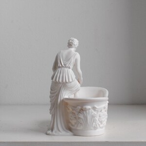 May include: A matte white figurine of a person in a draped garment standing beside a small, round basin. The basin features an ornate, raised relief pattern of acanthus leaves and floral motifs around its exterior, while the figure is posed.