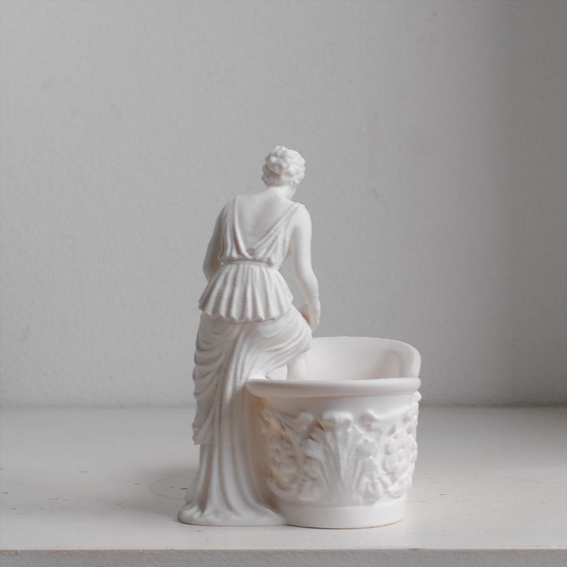 May include: A matte white figurine of a person in a draped garment standing beside a small, round basin. The basin features an ornate, raised relief pattern of acanthus leaves and floral motifs around its exterior, while the figure is posed.