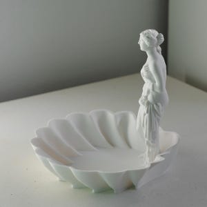 Puede incluir: Un cuenco decorativo de color blanco mate con forma de concha festoneada, que presenta una figura femenina cl&aacute;sica de pie unida a un lado. La escultura est&aacute; realizada en un material blanco s&oacute;lido y uniforme con l&iacute;neas suaves y un dise&ntilde;o limpio.