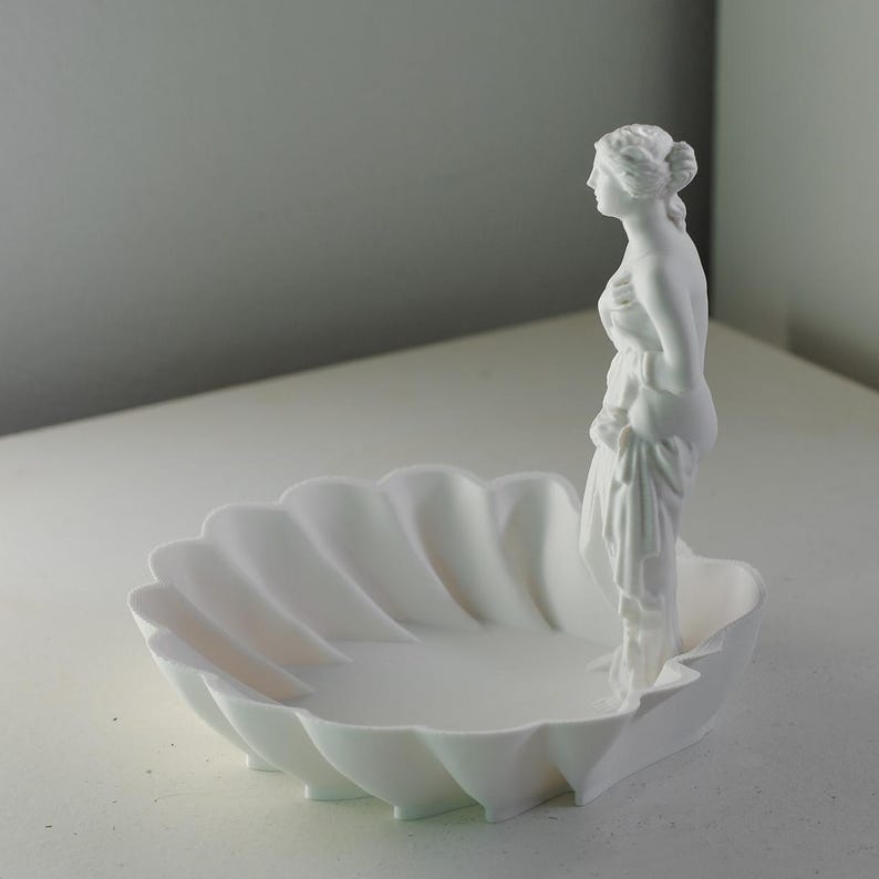 Puede incluir: Un cuenco decorativo de color blanco mate con forma de concha festoneada, que presenta una figura femenina cl&aacute;sica de pie unida a un lado. La escultura est&aacute; realizada en un material blanco s&oacute;lido y uniforme con l&iacute;neas suaves y un dise&ntilde;o limpio.