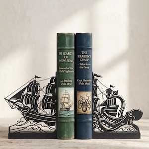 Puede incluir: Un par de sujetalibros de metal negro con un velero en un lado y un pulpo gigante en el otro, que sostienen dos libros de estilo antiguo titulados In Search of New Seas y The Kraken's Grasp, con detalladas ilustraciones n&aacute;uticas.