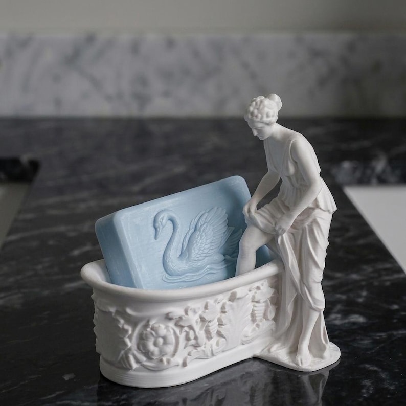 May include: A white ceramic soap dish shaped like a bathtub with ornate floral relief carvings. A standing figure in a draped garment is attached to the side. A rectangular light blue soap bar with a swan relief design rests inside the tub.
