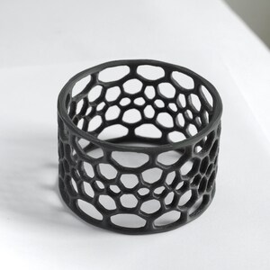 Puede incluir: Un objeto decorativo cil&iacute;ndrico negro con un patr&oacute;n de panal. El dise&ntilde;o calado presenta numerosas formas hexagonales irregulares, creando una textura visualmente interesante. El objeto probablemente est&aacute; hecho de un material s&oacute;lido y se encuentra sobre una superficie blanca.