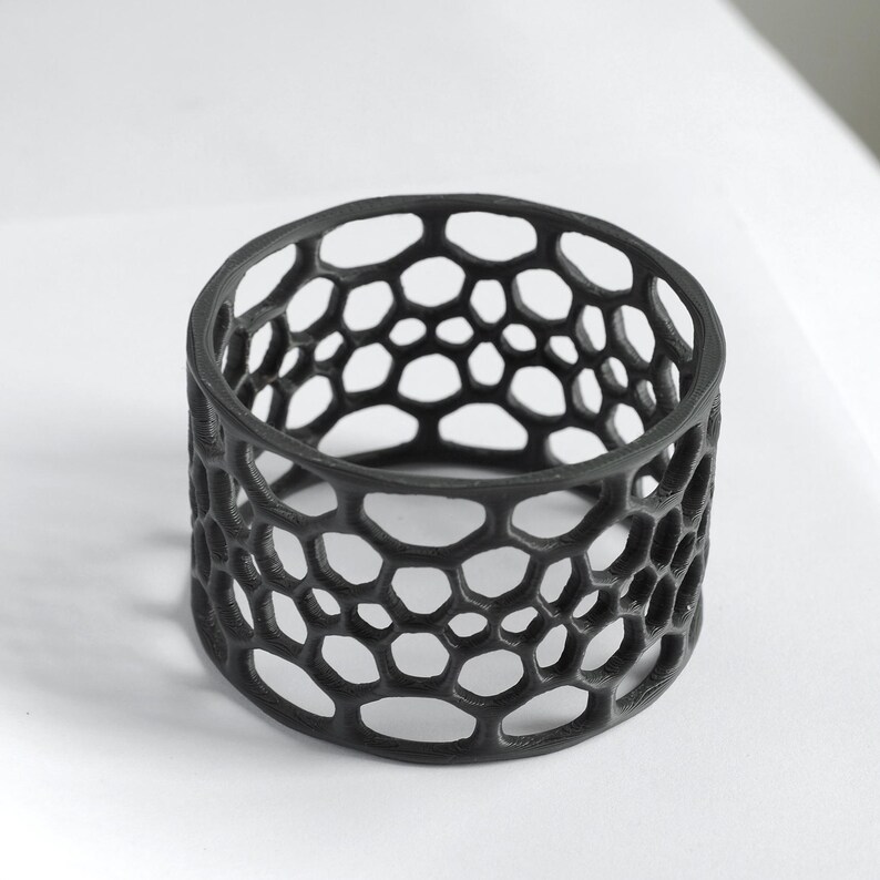 Puede incluir: Un objeto decorativo cil&iacute;ndrico negro con un patr&oacute;n de panal. El dise&ntilde;o calado presenta numerosas formas hexagonales irregulares, creando una textura visualmente interesante. El objeto probablemente est&aacute; hecho de un material s&oacute;lido y se encuentra sobre una superficie blanca.