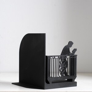 Puede incluir: Un sujetalibros de metal negro mate que presenta la silueta de una persona apoyada en una barandilla de balc&oacute;n decorativa mientras lee un libro. El dise&ntilde;o incluye un panel vertical s&oacute;lido y un recorte detallado de una figura.