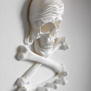 Puede incluir: Un perchero de pared blanco impreso en 3D que presenta una calavera con un pa&ntilde;uelo sobre dos huesos cruzados. La base de la estructura incorpora cuatro ganchos cil&iacute;ndricos que se extienden hacia afuera para colgar diversos objetos.