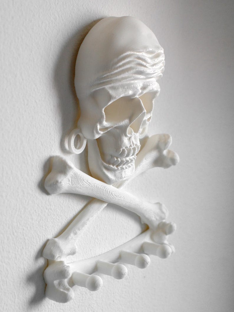 Puede incluir: Un perchero de pared blanco impreso en 3D que presenta una calavera con un pa&ntilde;uelo sobre dos huesos cruzados. La base de la estructura incorpora cuatro ganchos cil&iacute;ndricos que se extienden hacia afuera para colgar diversos objetos.