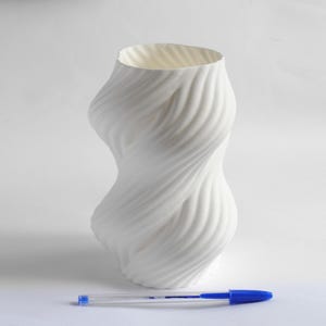 Puede incluir: Un jarr&oacute;n blanco con un dise&ntilde;o arremolinado texturizado. El jarr&oacute;n tiene una abertura ancha y una forma curva. Un bol&iacute;grafo azul y transparente est&aacute; en primer plano. El jarr&oacute;n mide aproximadamente 20 cm de alto.