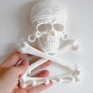 Puede incluir: Un perchero de pared impreso en 3D en color blanco mate con forma de cr&aacute;neo humano, un pa&ntilde;uelo y dos huesos cruzados. El cr&aacute;neo presenta dilatadores circulares en las orejas y el hueso inferior incluye cuatro peque&ntilde;os ganchos redondeados.