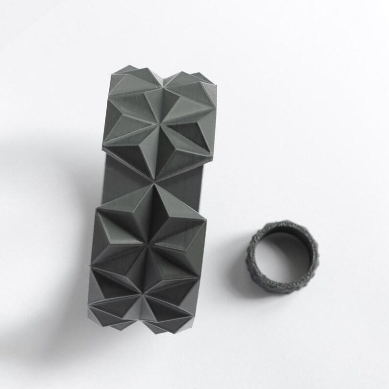 May include: A dark gray geometric bracelet and ring set. The bracelet features a repeating starburst pattern, while the ring has a textured, ridged design. Both items are made of a matte material and sit on a white surface.