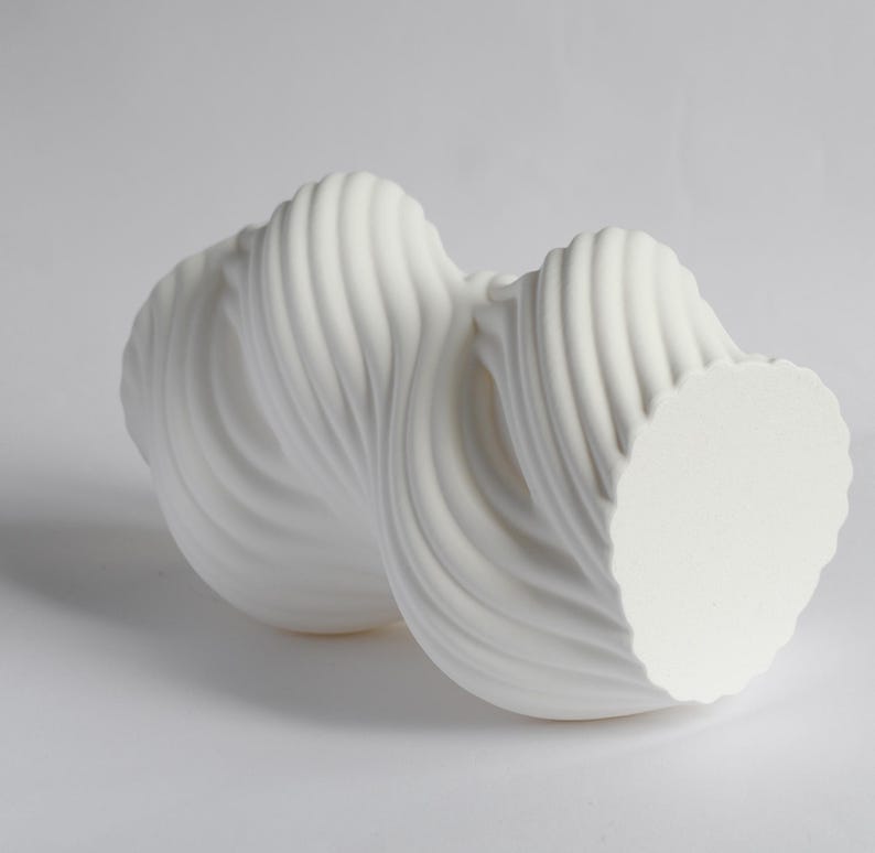 Puede incluir: Una escultura abstracta blanca con un dise&ntilde;o fluido y retorcido. La escultura tiene una superficie texturizada y acanalada y una base circular. La forma general recuerda a una ola o una cinta. La pieza est&aacute; hecha de un material s&oacute;lido y opaco.