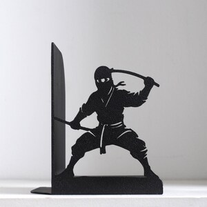 Puede incluir: Un sujetalibros de metal negro con la silueta de un ninja en posici&oacute;n agachada. La figura sostiene una espada sobre su cabeza con una mano y extiende una segunda espada hacia afuera con la otra. El dise&ntilde;o est&aacute; cortado en una pieza de metal.