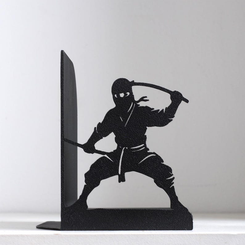 Puede incluir: Un sujetalibros de metal negro con la silueta de un ninja en posici&oacute;n agachada. La figura sostiene una espada sobre su cabeza con una mano y extiende una segunda espada hacia afuera con la otra. El dise&ntilde;o est&aacute; cortado en una pieza de metal.