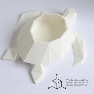 Puede incluir: Una maceta blanca con forma de tortuga geom&eacute;trica. La maceta mide 16,1 cm de ancho, 4,4 cm de alto y 19 cm de profundidad. El dise&ntilde;o presenta una forma poligonal con una abertura central.
