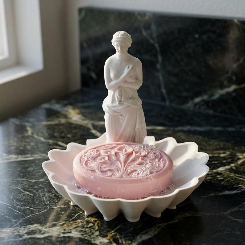 Puede incluir: Una jabonera blanca con forma de concha festoneada que presenta una escultura central de una figura femenina de pie. Una pastilla de jab&oacute;n redonda de color rosa claro con un patr&oacute;n floral en relieve de calas descansa en el plato frente a la figura.