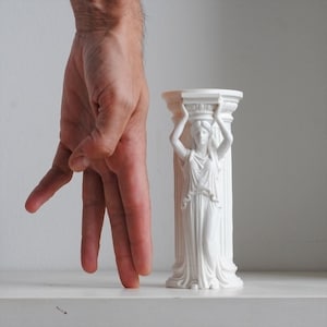 Puede incluir: Un portavelas cil&iacute;ndrico blanco con forma de columna que presenta una figura de cari&aacute;tide esculpida con una t&uacute;nica drapeada. La figura tiene los brazos levantados para sostener el capitel superior. El objeto tiene un acabado blanco mate.