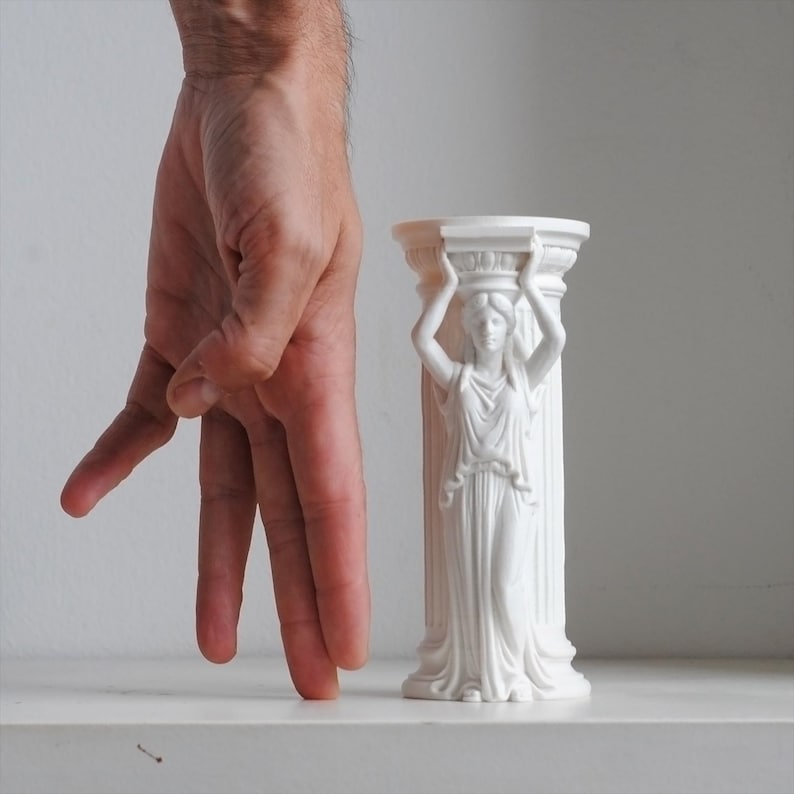 Puede incluir: Un portavelas cil&iacute;ndrico blanco con forma de columna que presenta una figura de cari&aacute;tide esculpida con una t&uacute;nica drapeada. La figura tiene los brazos levantados para sostener el capitel superior. El objeto tiene un acabado blanco mate.