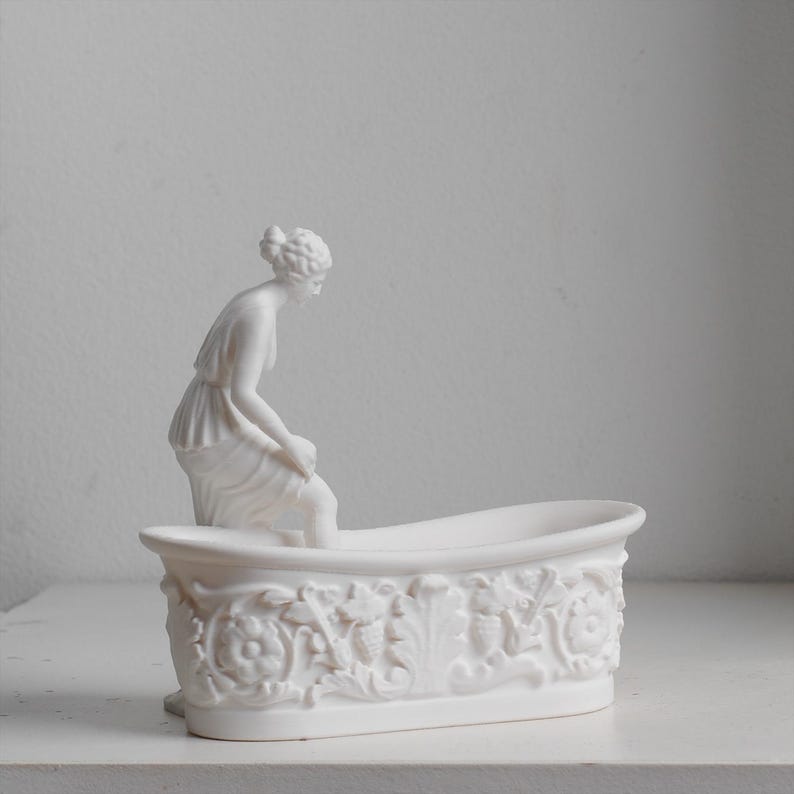 May include: A white matte ceramic figurine of a person in a draped garment sitting on the edge of a bathtub. The exterior of the bathtub features a detailed relief pattern of floral and leaf motifs. The entire object is a solid, uniform white.