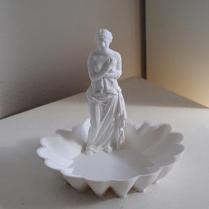 Puede incluir: Un plato decorativo de color blanco mate que presenta una figura escult&oacute;rica central de una mujer cl&aacute;sica envuelta en tela. La base del plato tiene un borde festoneado similar a un p&eacute;talo y todo el objeto tiene un acabado blanco uniforme.