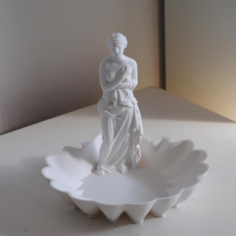 Puede incluir: Un plato decorativo de color blanco mate que presenta una figura escult&oacute;rica central de una mujer cl&aacute;sica envuelta en tela. La base del plato tiene un borde festoneado similar a un p&eacute;talo y todo el objeto tiene un acabado blanco uniforme.
