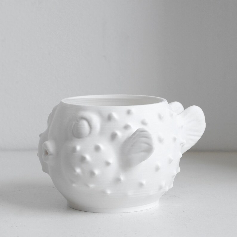 Puede incluir: Una taza de cer&aacute;mica blanca mate con forma de pez globo. El recipiente presenta un cuerpo redondeado con espinas texturizadas en relieve por toda la superficie, una boca peque&ntilde;a y saliente, cuencas oculares definidas y un asa en forma de aleta.
