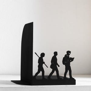May include: A black metal bookend featuring a silhouette design of three figures walking in a line. The first figure carries a staff, the second walks with a bag, and the third figure holds an open book while wearing a backpack.