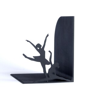 May include: Black metal bookends featuring silhouettes of two ballerinas in graceful dance poses. One ballerina is leaping, while the other is seated in a split. The bookends are designed to hold books upright on a shelf.