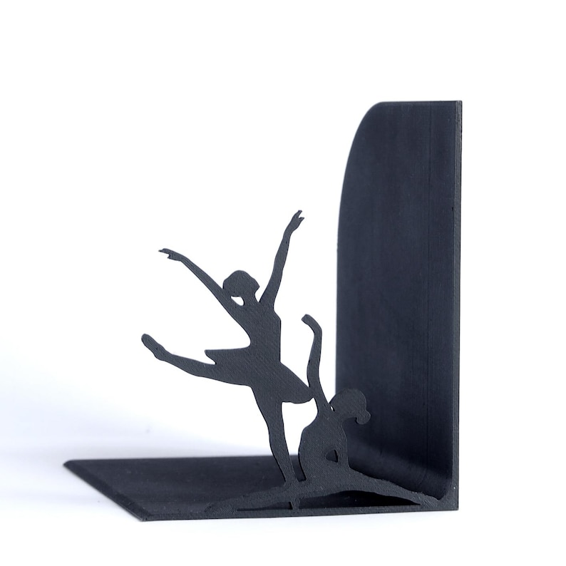 May include: Black metal bookends featuring silhouettes of two ballerinas in graceful dance poses. One ballerina is leaping, while the other is seated in a split. The bookends are designed to hold books upright on a shelf.