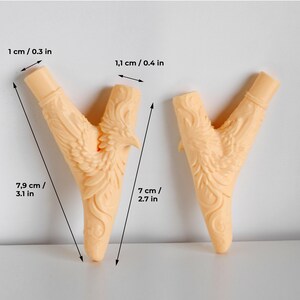 Puede incluir: Dos objetos en forma de Y de color melocot&oacute;n claro con un relieve tallado de un p&aacute;jaro con las alas extendidas. El objeto de la izquierda mide 7,9 cm de largo total, 1 cm en la parte superior y 1,1 cm en la rama. El de la derecha mide 7 cm.