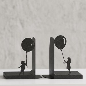 May include: A pair of black metal bookends featuring a silhouette design. One bookend depicts a child holding a balloon, while the other shows a person holding a balloon. The metal has a textured finish and a sturdy L-shaped base for support.