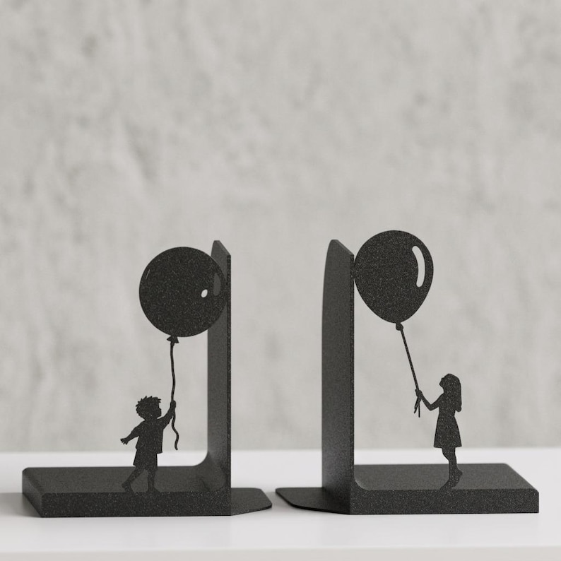 May include: A pair of black metal bookends featuring a silhouette design. One bookend depicts a child holding a balloon, while the other shows a person holding a balloon. The metal has a textured finish and a sturdy L-shaped base for support.