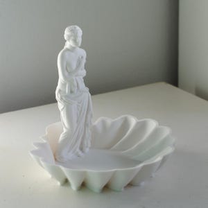 Puede incluir: Una bandeja decorativa de color blanco mate que presenta la figura esculpida de una persona de pie envuelta en tela, situada dentro de un cuenco con forma de concha festoneada. El objeto es monocrom&aacute;tico y tiene l&iacute;neas suaves.