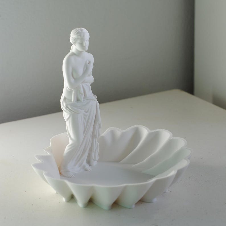 Puede incluir: Una bandeja decorativa de color blanco mate que presenta la figura esculpida de una persona de pie envuelta en tela, situada dentro de un cuenco con forma de concha festoneada. El objeto es monocrom&aacute;tico y tiene l&iacute;neas suaves.