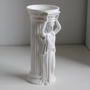 Puede incluir: Un florero cil&iacute;ndrico de acabado blanco mate que presenta una figura de cari&aacute;tide esculpida integrada en el lateral. La figura se muestra con los brazos levantados, sosteniendo el borde superior de la estructura de columna estriada, similar a un elemento cl&aacute;sico.