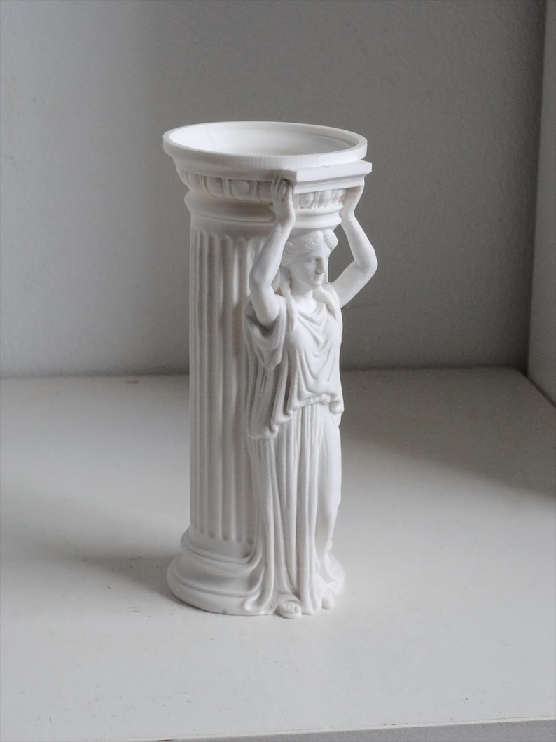 Puede incluir: Un florero cil&iacute;ndrico de acabado blanco mate que presenta una figura de cari&aacute;tide esculpida integrada en el lateral. La figura se muestra con los brazos levantados, sosteniendo el borde superior de la estructura de columna estriada, similar a un elemento cl&aacute;sico.