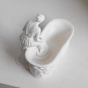 May include: A matte white ceramic sculpture featuring a figure in a draped garment leaning over a small, oval-shaped bathtub. The exterior of the tub is decorated with embossed floral and scroll patterns, while the figure is positioned at the edge.