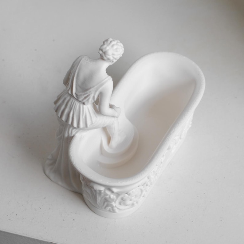 May include: A matte white ceramic sculpture featuring a figure in a draped garment leaning over a small, oval-shaped bathtub. The exterior of the tub is decorated with embossed floral and scroll patterns, while the figure is positioned at the edge.