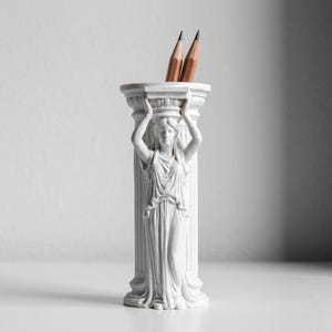 Puede incluir: Un portal&aacute;pices cil&iacute;ndrico blanco con forma de columna cl&aacute;sica que presenta la figura esculpida de una persona sosteniendo el borde superior. La figura viste ropa drapeada y est&aacute; de pie junto a la columna. Dos l&aacute;pices de madera est&aacute;n dentro.