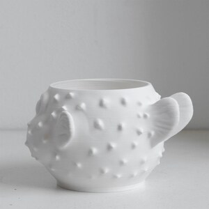Puede incluir: Una taza de cer&aacute;mica blanca mate con forma de pez globo, que presenta un cuerpo redondeado cubierto de peque&ntilde;as p&uacute;as en relieve, una amplia abertura circular en la parte superior y una aleta prominente en el lateral que sirve de asa.