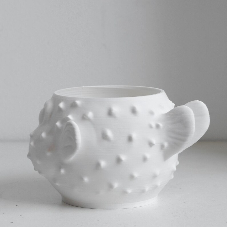 Puede incluir: Una taza de cer&aacute;mica blanca mate con forma de pez globo, que presenta un cuerpo redondeado cubierto de peque&ntilde;as p&uacute;as en relieve, una amplia abertura circular en la parte superior y una aleta prominente en el lateral que sirve de asa.