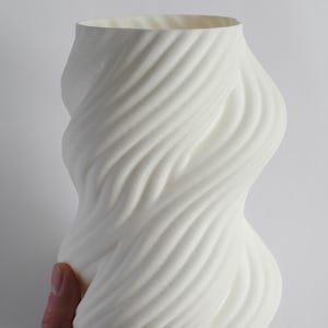 Puede incluir: Un jarr&oacute;n de cer&aacute;mica blanco con un dise&ntilde;o arremolinado y texturizado. El jarr&oacute;n tiene una abertura ancha y una forma org&aacute;nica curvada. La superficie presenta l&iacute;neas diagonales en relieve que crean un efecto espiral. El jarr&oacute;n es sostenido por una mano.
