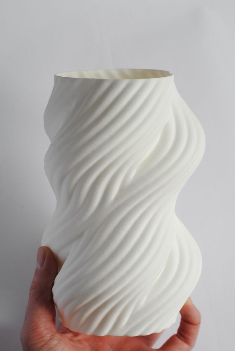 Puede incluir: Un jarr&oacute;n de cer&aacute;mica blanco con un dise&ntilde;o arremolinado y texturizado. El jarr&oacute;n tiene una abertura ancha y una forma org&aacute;nica curvada. La superficie presenta l&iacute;neas diagonales en relieve que crean un efecto espiral. El jarr&oacute;n es sostenido por una mano.
