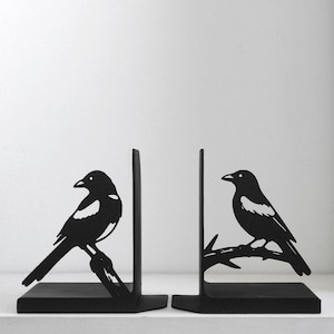 Puede incluir: Un par de sujetalibros de metal negro con un dise&ntilde;o de silueta de una urraca posada en una rama. Cada sujetalibros tiene una base plana y una placa de soporte vertical, con la figura del ave extendi&eacute;ndose desde el lado del panel met&aacute;lico.