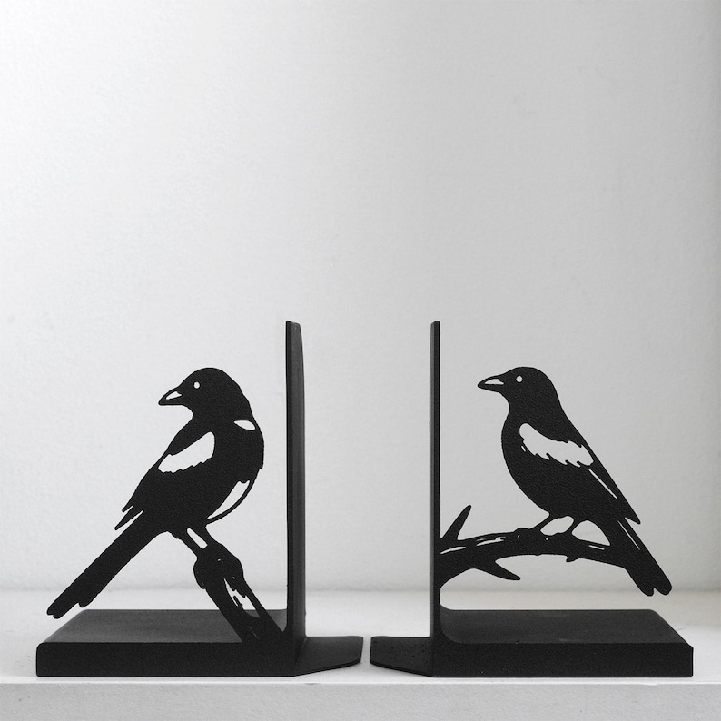 Puede incluir: Un par de sujetalibros de metal negro con un dise&ntilde;o de silueta de una urraca posada en una rama. Cada sujetalibros tiene una base plana y una placa de soporte vertical, con la figura del ave extendi&eacute;ndose desde el lado del panel met&aacute;lico.