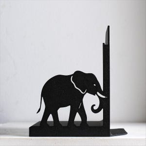 Puede incluir: Un sujetalibros de metal negro con la silueta de un elefante caminando hacia un panel de soporte vertical. El dise&ntilde;o est&aacute; cortado de una sola pieza de metal oscuro y texturizado, mostrando el perfil, la trompa y la oreja del elefante.