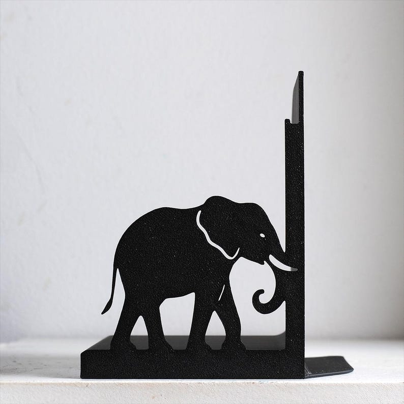 Puede incluir: Un sujetalibros de metal negro con la silueta de un elefante caminando hacia un panel de soporte vertical. El dise&ntilde;o est&aacute; cortado de una sola pieza de metal oscuro y texturizado, mostrando el perfil, la trompa y la oreja del elefante.