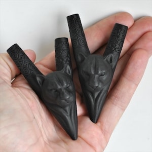 Puede incluir: Dos objetos de color negro mate en forma de V que presentan una cabeza de pantera esculpida en el centro y patrones intrincados y arremolinados grabados a lo largo de los dos tallos superiores. Tienen un acabado oscuro y una base puntiaguda.